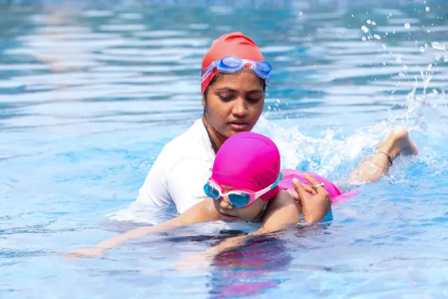 Une monitrice guide un enfant portant un bonnet et des lunettes dans une piscine extérieure.