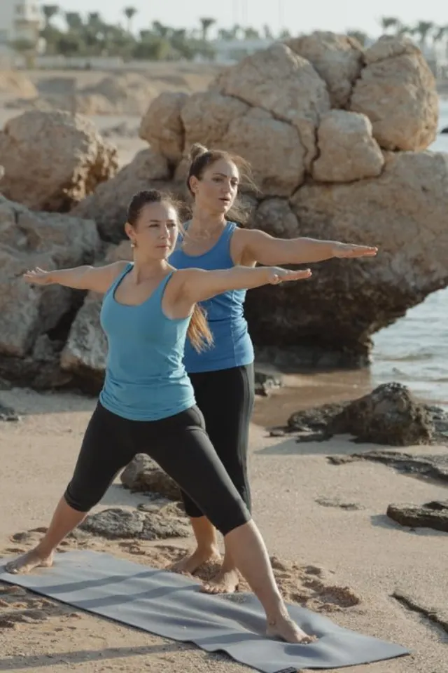 Deux femmes pratiquent le yoga sur une plage, l'une accompagnant la posture de l'autre.